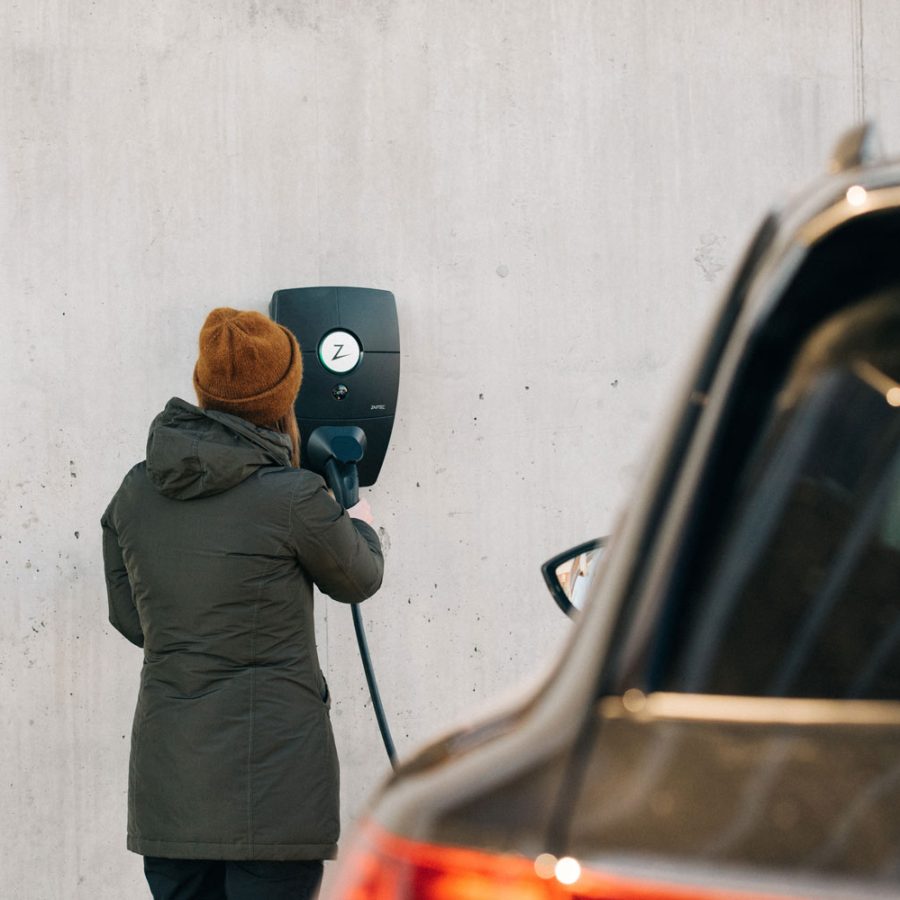 Dame i grønn vinterjakke og oransje lader elbil fra en sort moderne ladeboks montert på betongvegg.